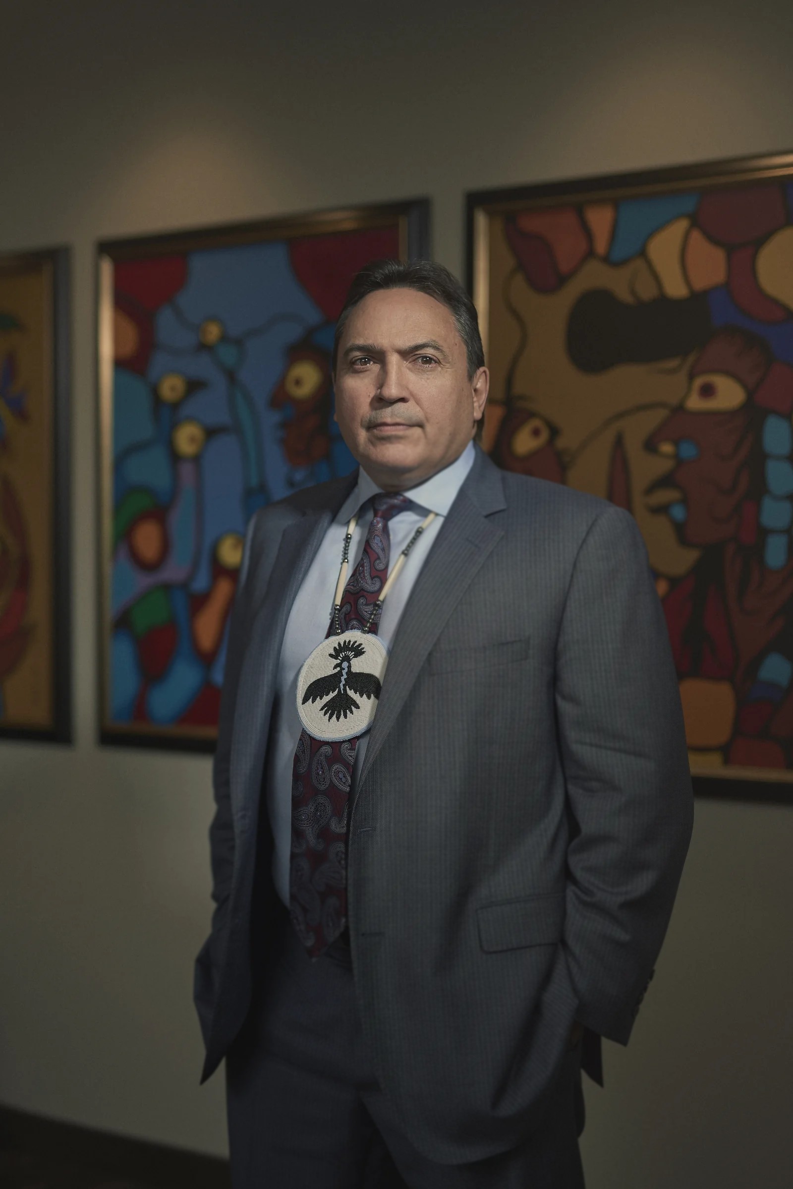 Perry Bellegarde in a grey suit and beaded thunderbird medallion, standing before Woodland-style paintings